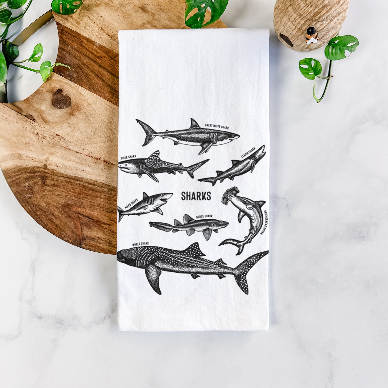 White cotton tea towel printed with a chart of black ink shark illustrations, including a whale shark, hammerhead and great white, on a marble counter beside a round wooden cutting board and greenery.