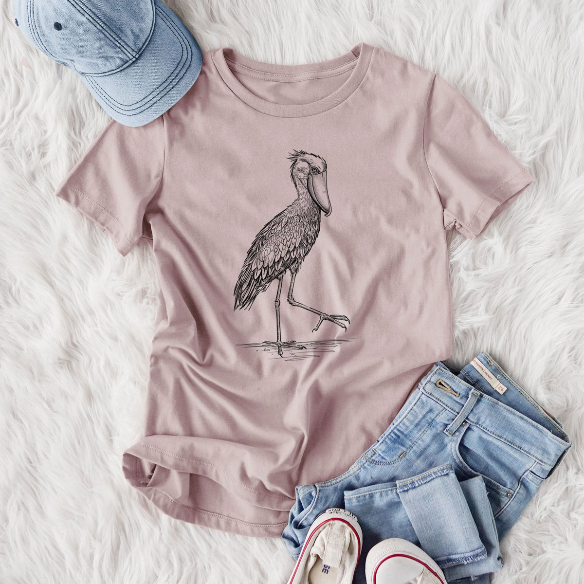 Women's relaxed fit cotton t-shirt printed with a black ink illustration of a shoebill stork standing on one leg, styled flat on a white faux fur rug with jeans.