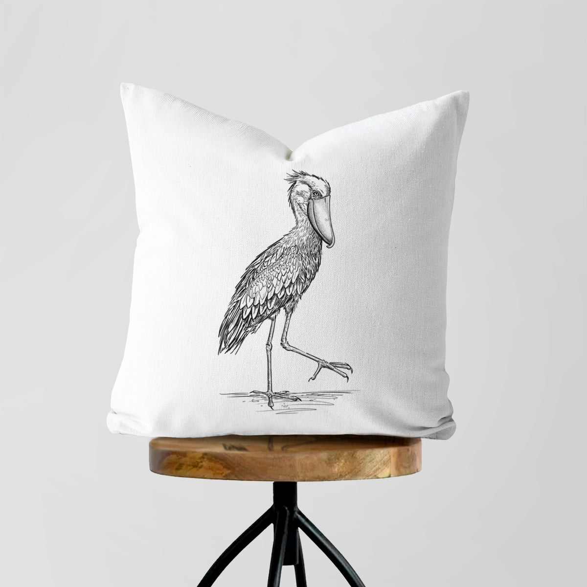 White pillow cover printed with a black ink illustration of a shoebill stork standing on one leg, on a wooden stool.
