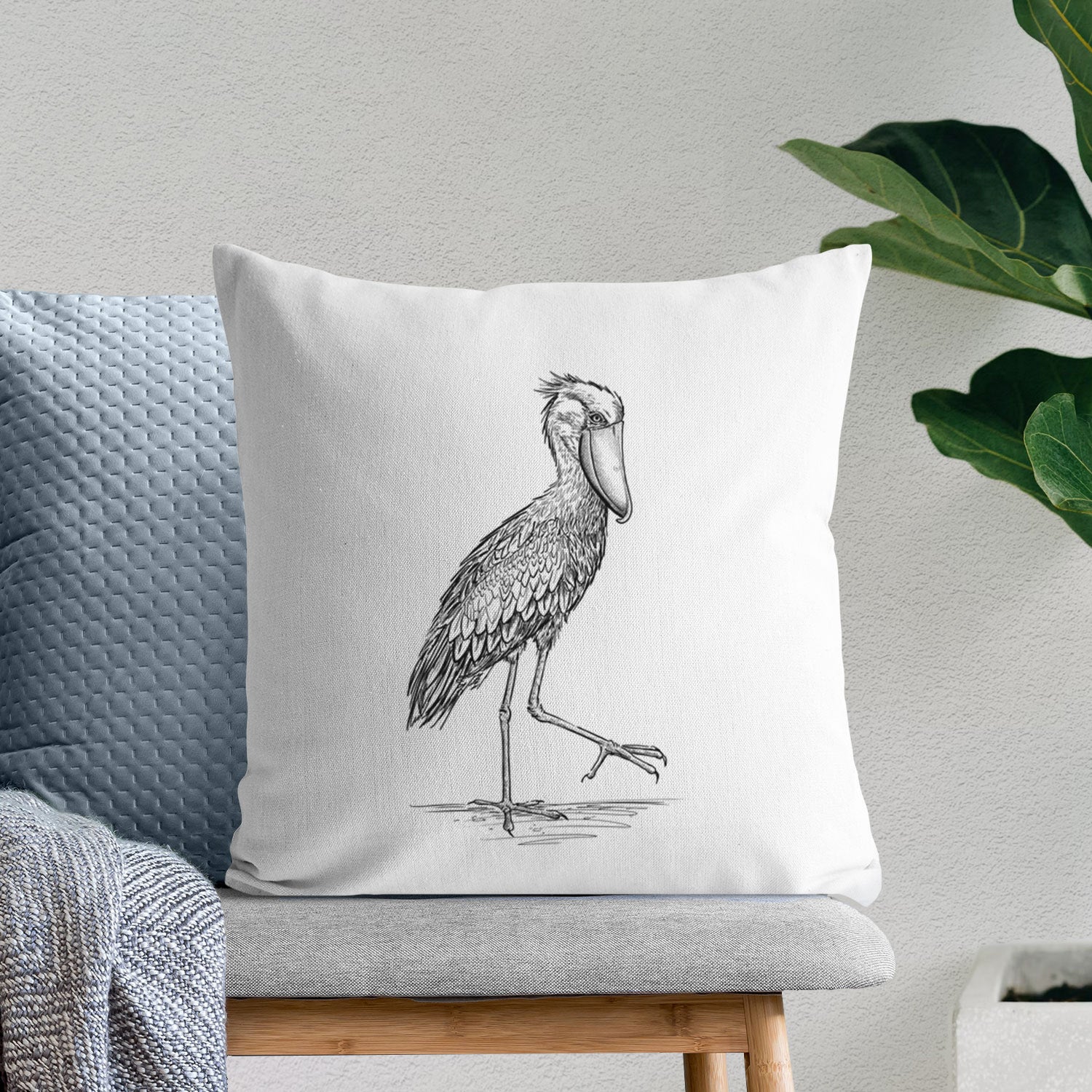 White pillow cover printed with a black ink illustration of a shoebill stork standing on one leg, on a bench beside plants.