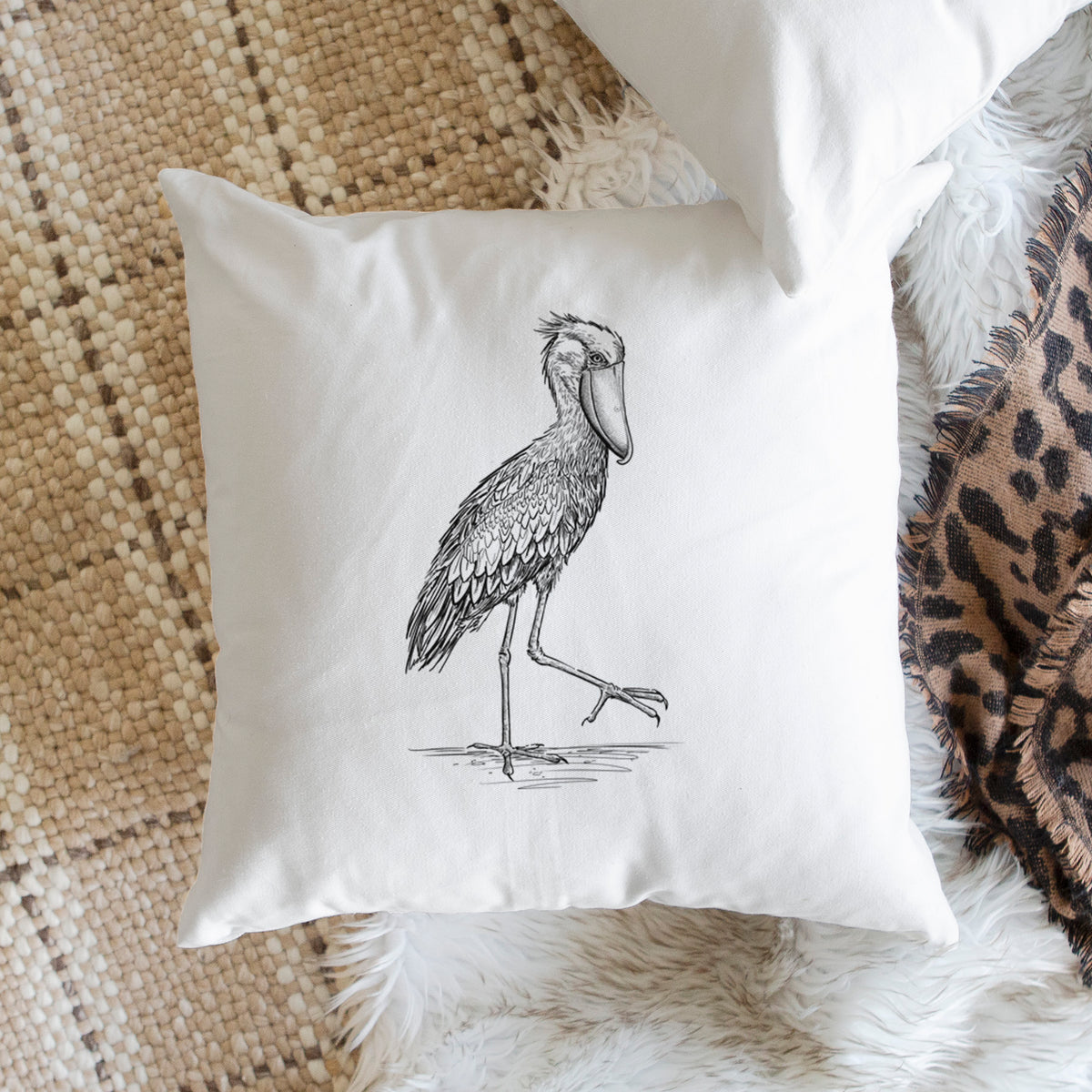 White pillow cover printed with a black ink illustration of a shoebill stork standing on one leg, on a woven chair.