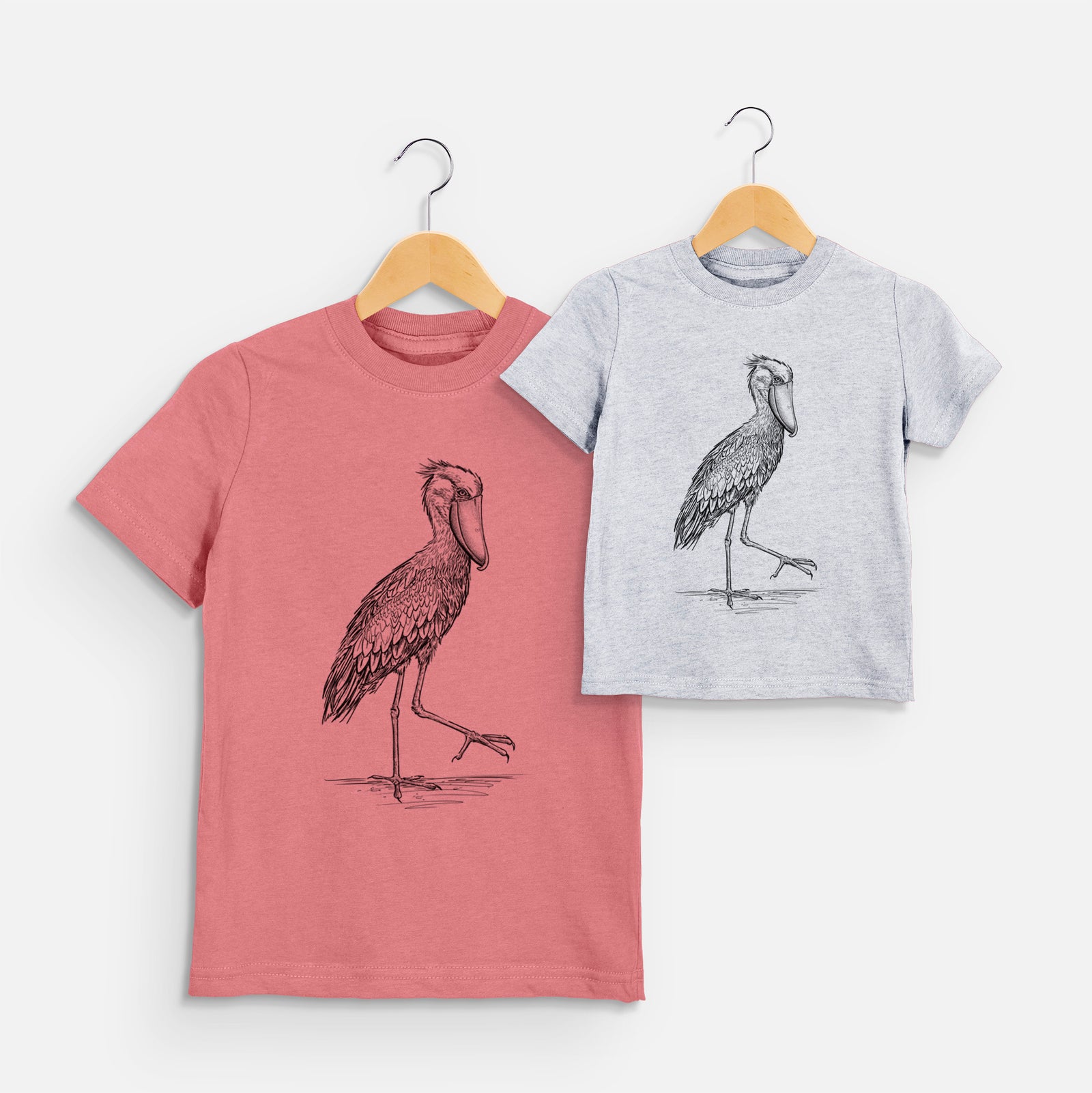 Two youth t-shirts in different colors on hangers, printed with a black ink illustration of a shoebill stork standing on one leg.