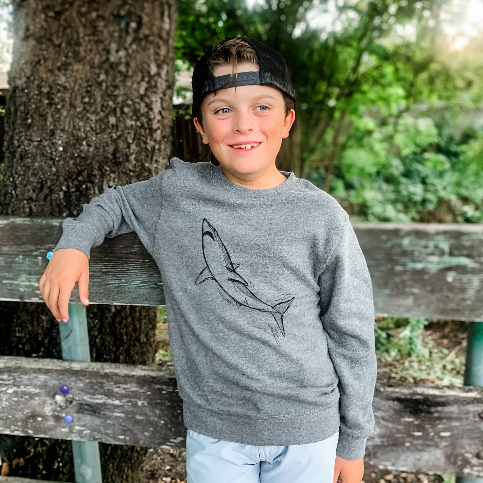 Boy wearing a youth crewneck sweatshirt printed with a black ink illustration of a shortfin mako shark diving, outdoors.