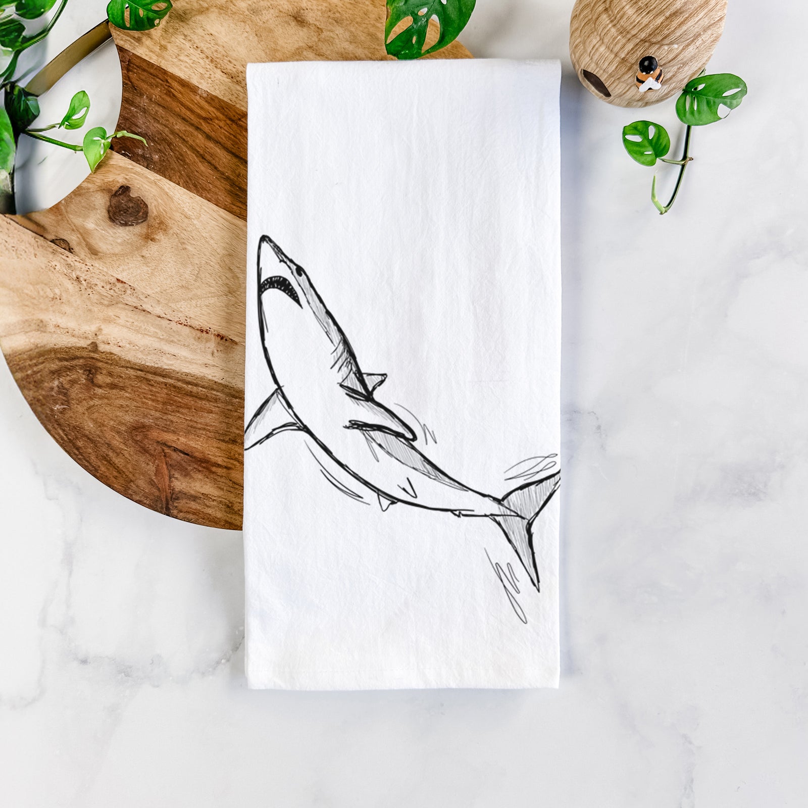 White cotton tea towel printed with a black ink illustration of a shortfin mako shark diving, on a marble counter beside a round wooden cutting board and greenery.