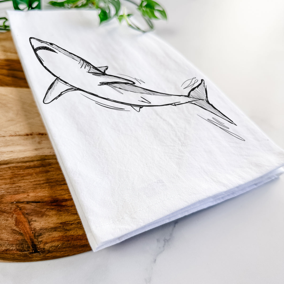 Close-up of a folded white tea towel on a wooden board, showing a black ink illustration of a shortfin mako shark diving.