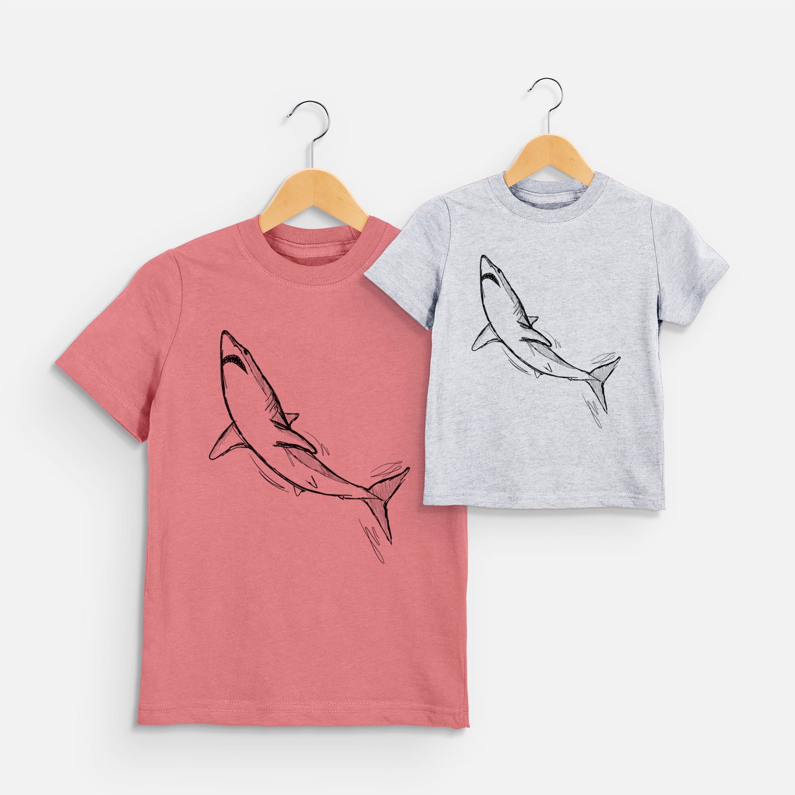 Two youth t-shirts in different colors on hangers, printed with a black ink illustration of a shortfin mako shark diving.