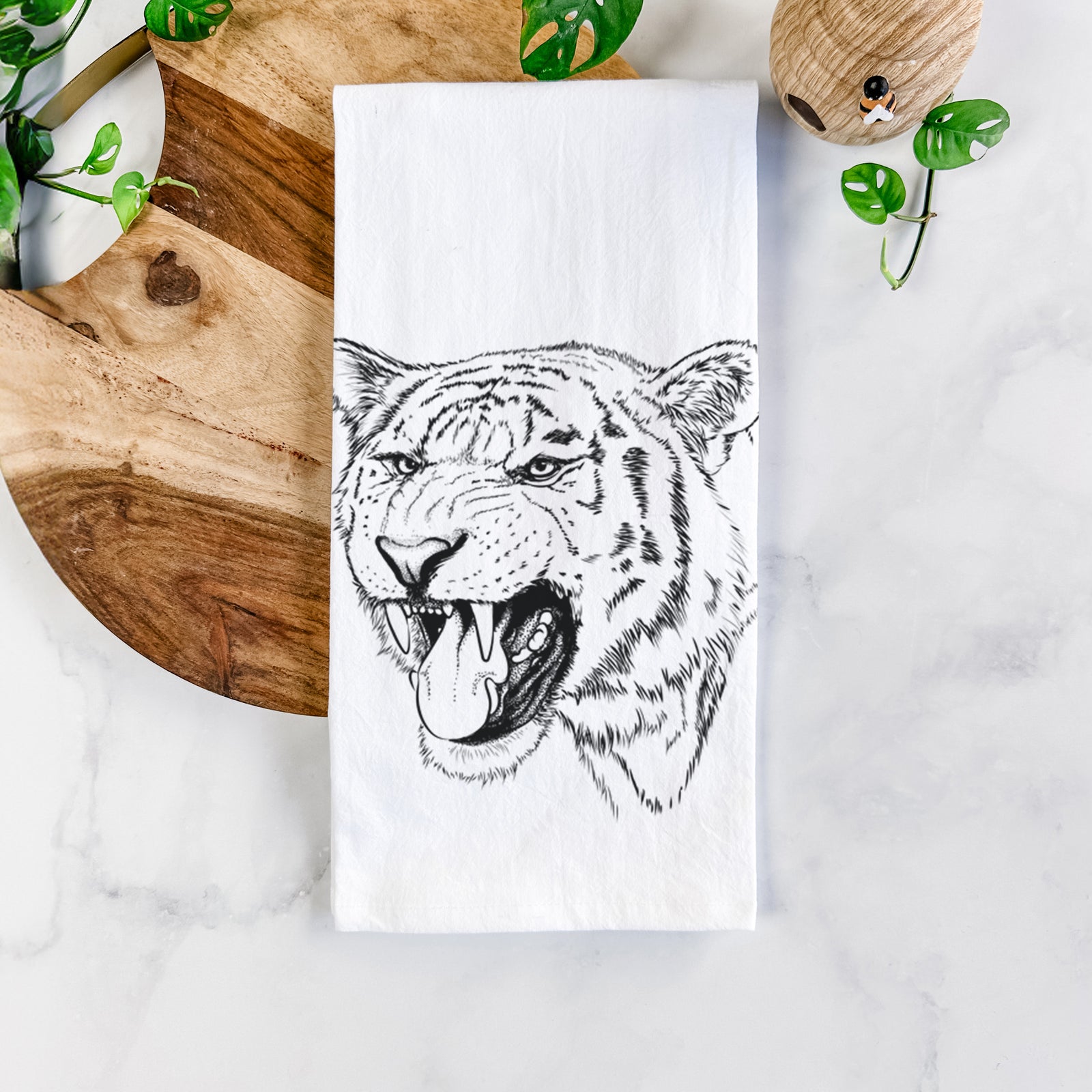 White cotton tea towel printed with a detailed black ink portrait of a roaring Siberian tiger, on a marble counter beside a round wooden cutting board and greenery.