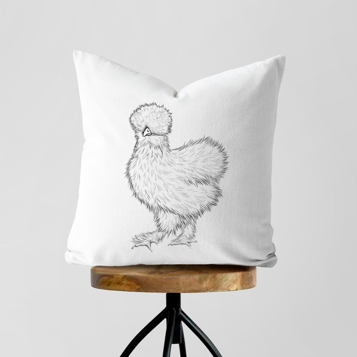 White pillow cover printed with a black ink illustration of a fluffy silkie chicken, on a wooden stool.