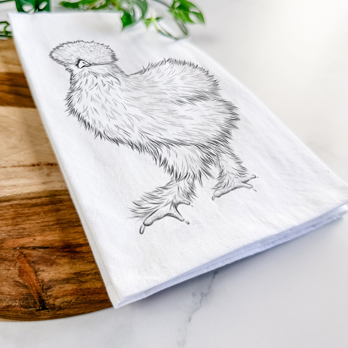 Close-up of a folded white tea towel on a wooden board, showing a black ink illustration of a fluffy silkie chicken.