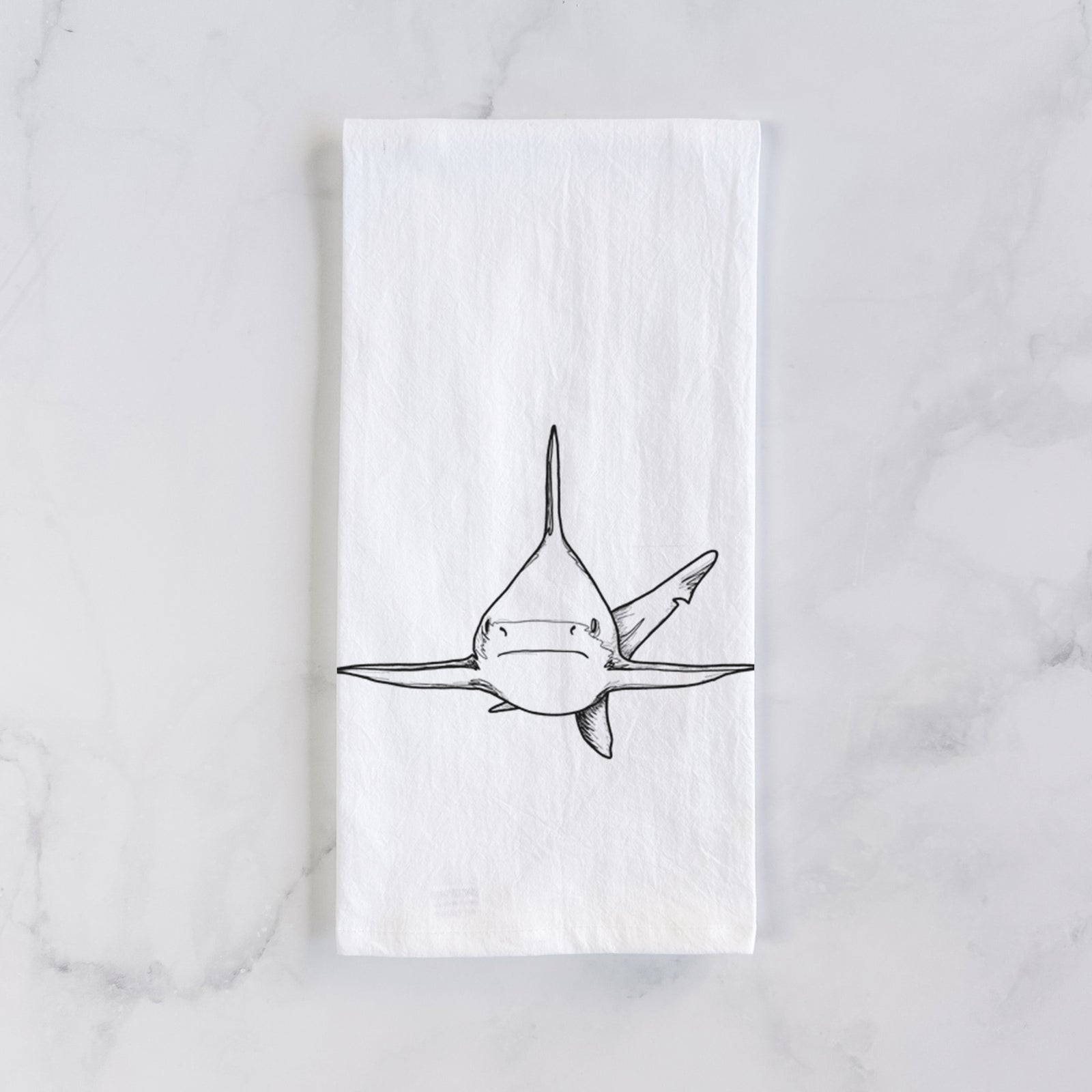 White cotton tea towel printed with a black ink illustration of a silvertip shark swimming head-on, on a marble counter beside a round wooden cutting board and greenery.