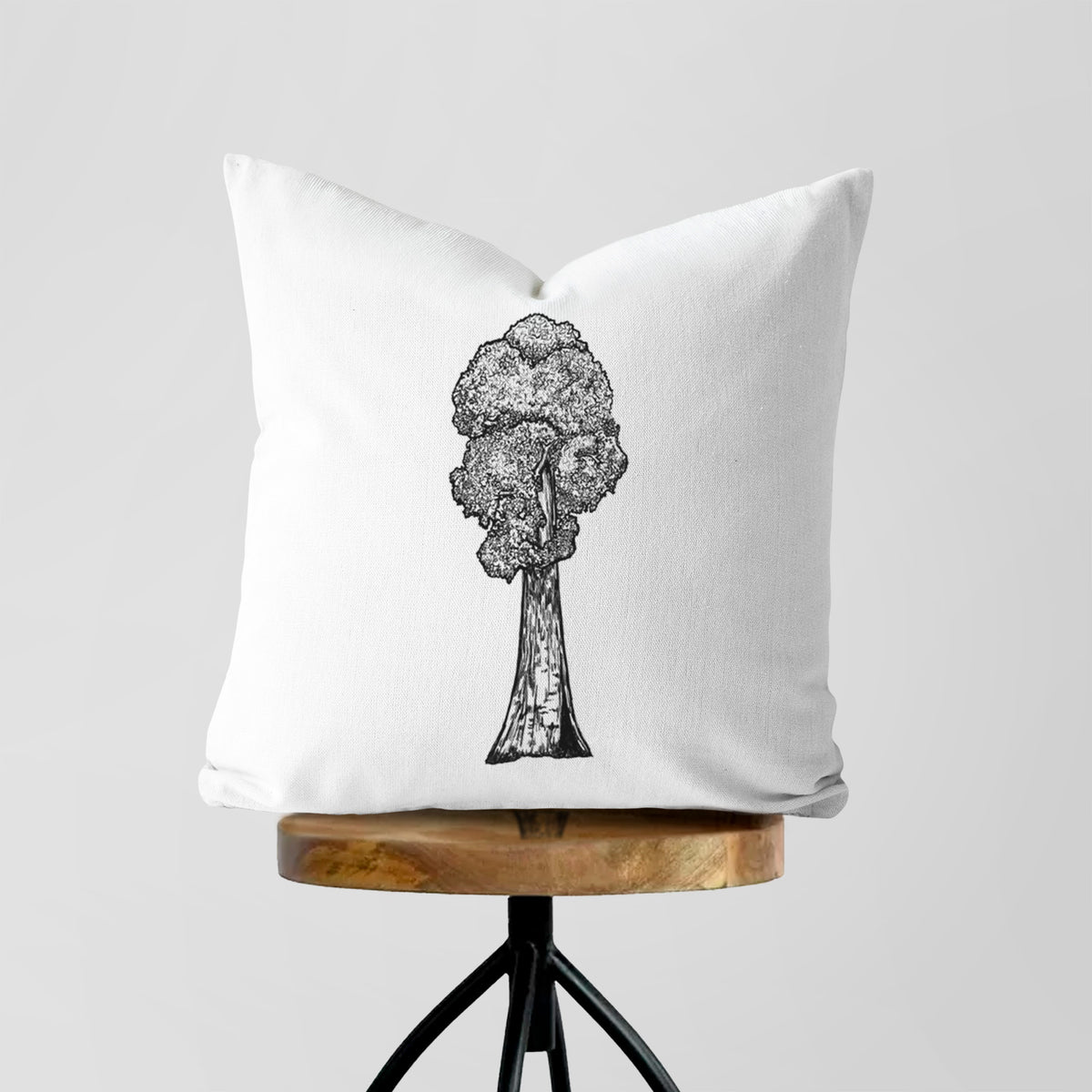 White pillow cover printed with a black ink illustration of a single giant sequoia tree, on a wooden stool.