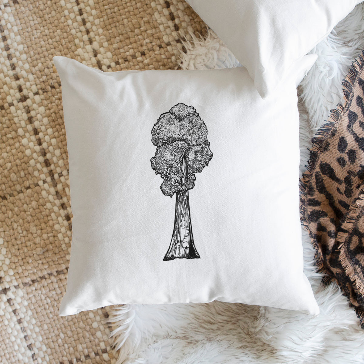White pillow cover printed with a black ink illustration of a single giant sequoia tree, on a woven chair.