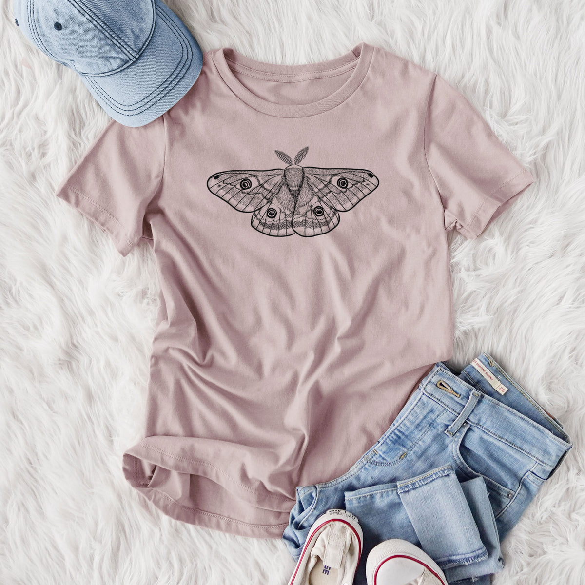 Women's relaxed fit cotton t-shirt printed with a detailed black ink illustration of a small emperor moth with eyespots on its wings, styled flat on a white faux fur rug with jeans.