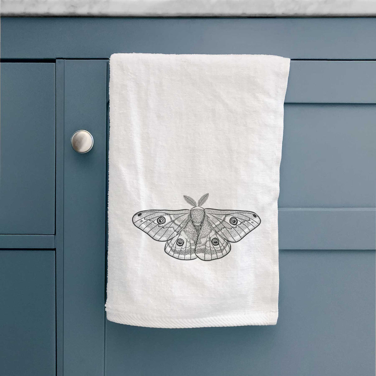 White hand towel printed with a detailed black ink illustration of a small emperor moth with eyespots on its wings, hanging on a cabinet door.