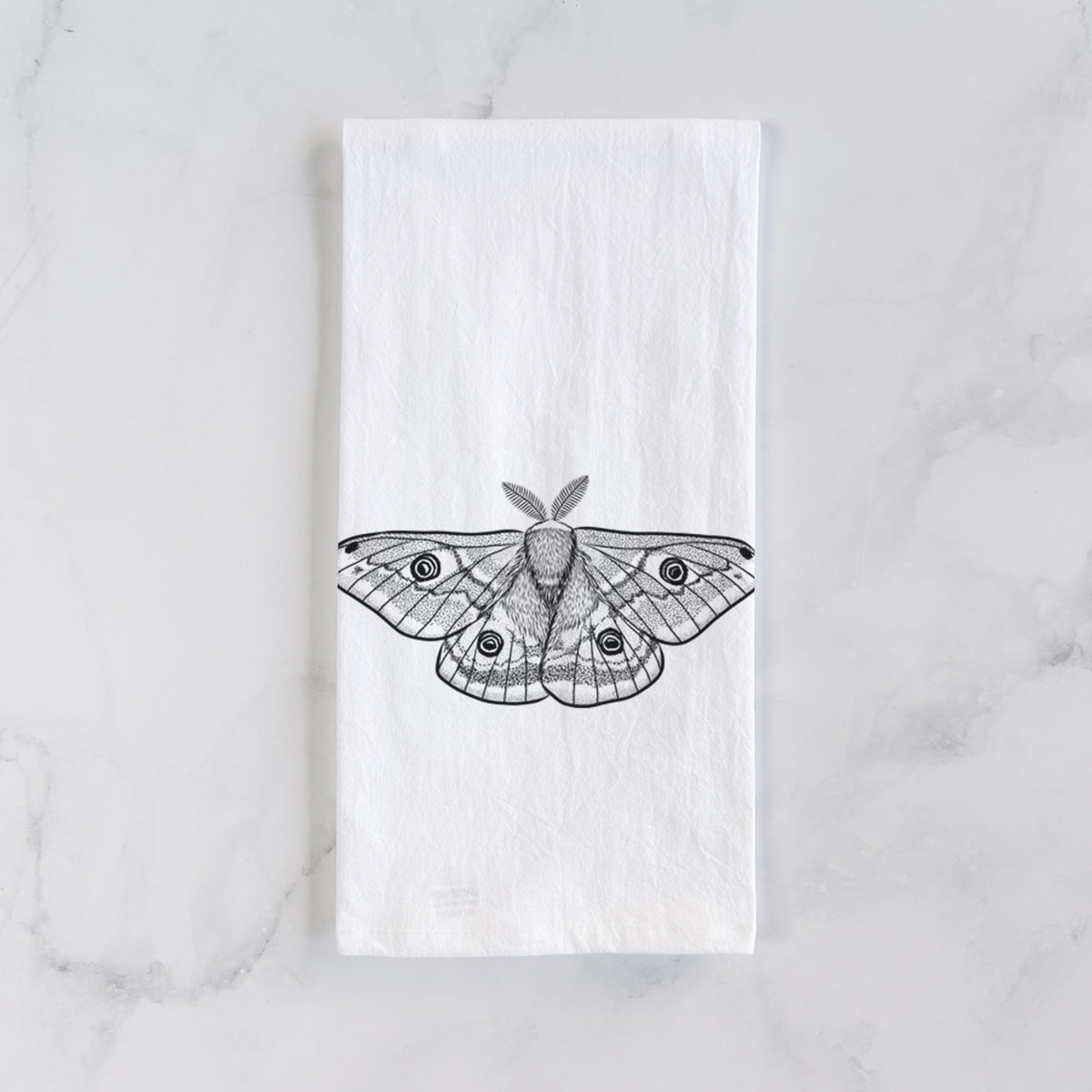 White cotton tea towel laid flat on marble, printed with a detailed black ink illustration of a small emperor moth with eyespots on its wings.