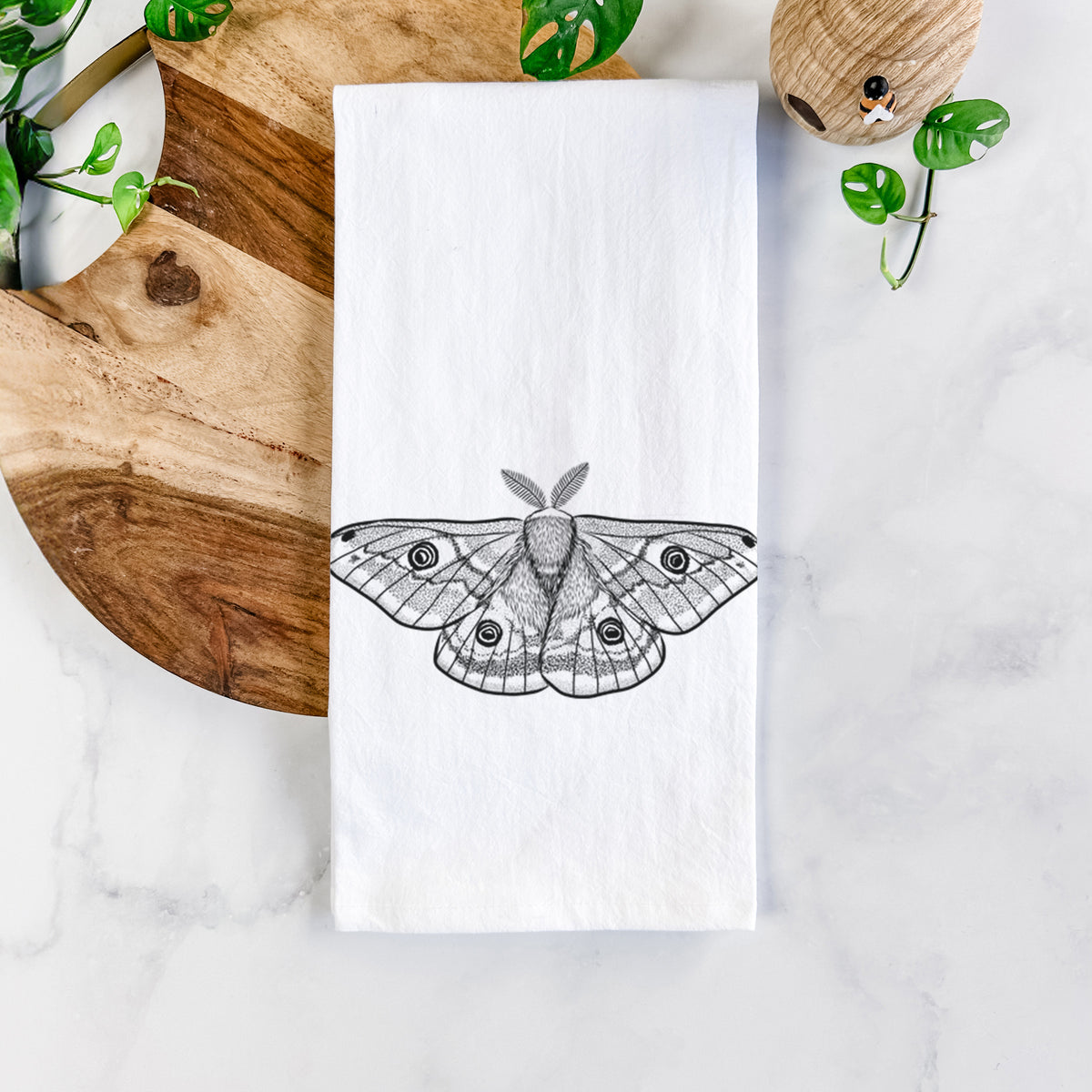 White cotton tea towel printed with a detailed black ink illustration of a small emperor moth with eyespots on its wings, on a marble counter beside a round wooden cutting board and greenery.