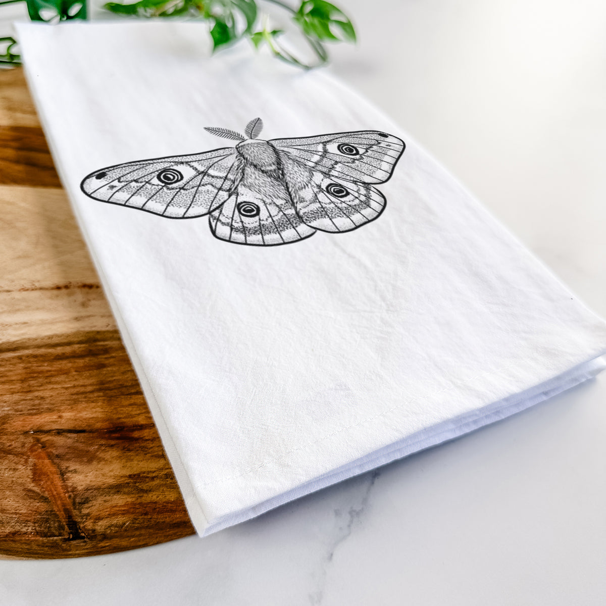 Close-up of a folded white tea towel on a wooden board, showing a detailed black ink illustration of a small emperor moth with eyespots on its wings.