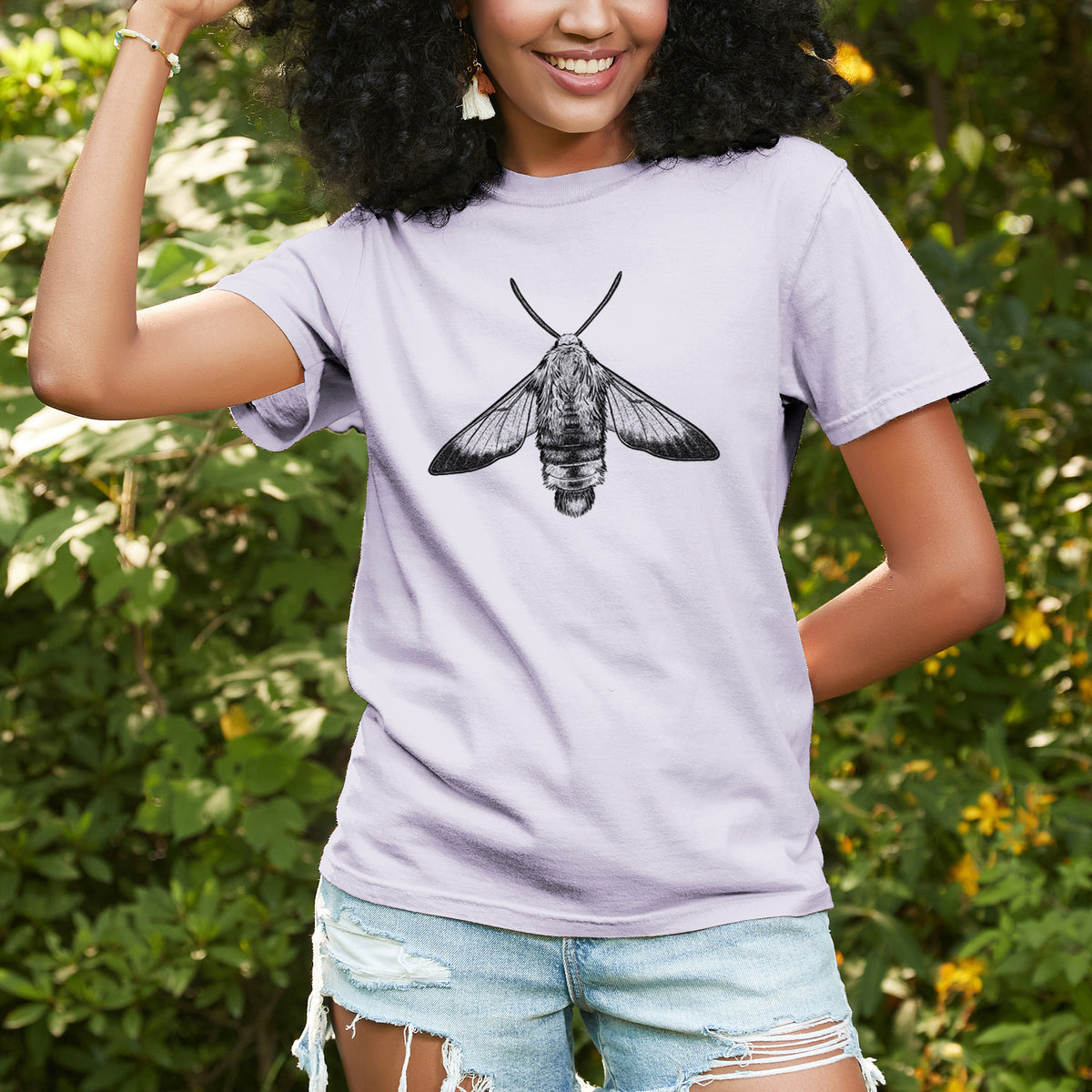 Woman wearing a Comfort Colors t-shirt printed with a detailed black ink illustration of a snowberry clearwing moth, seen from above, outdoors.