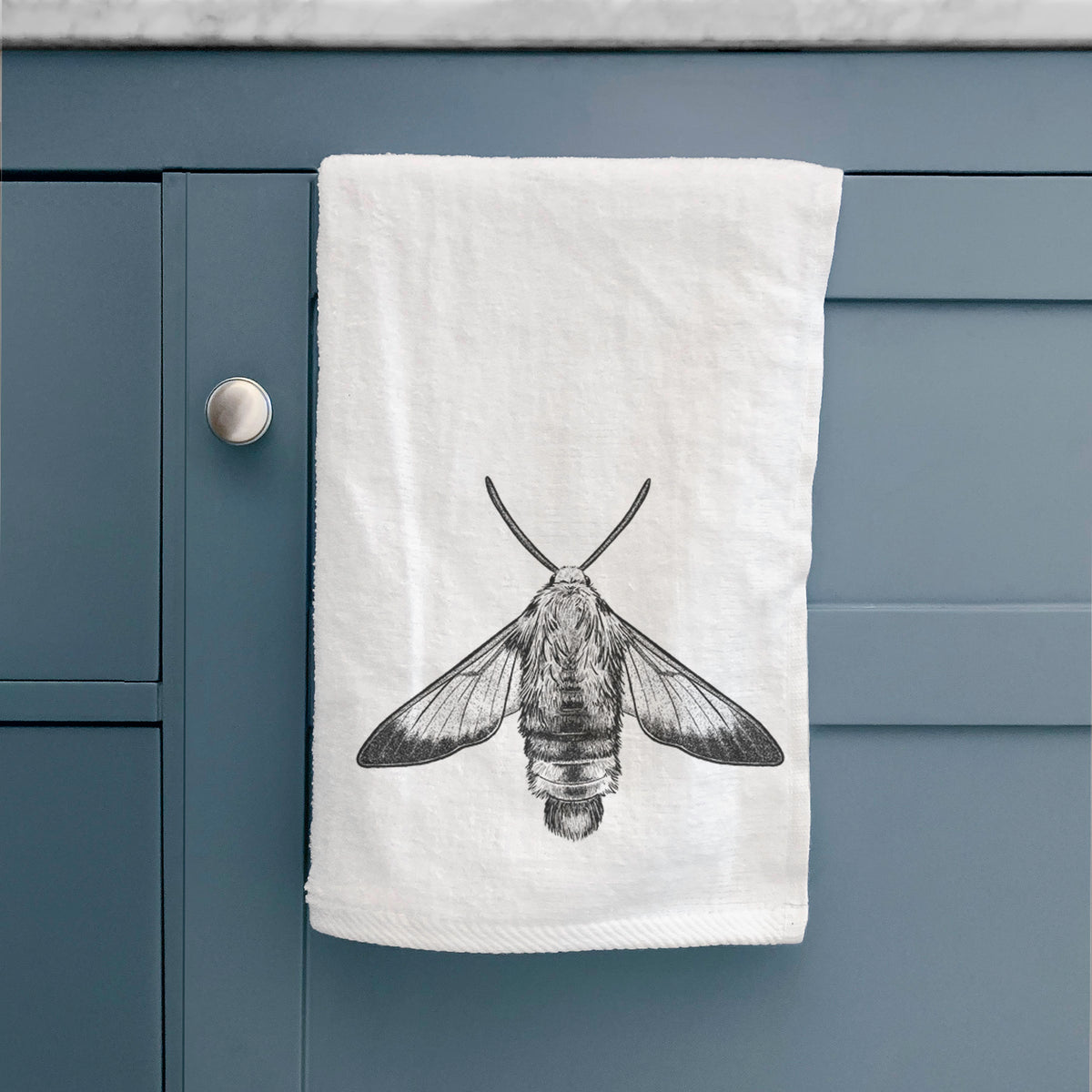 White hand towel printed with a detailed black ink illustration of a snowberry clearwing moth, seen from above, hanging on a cabinet door.