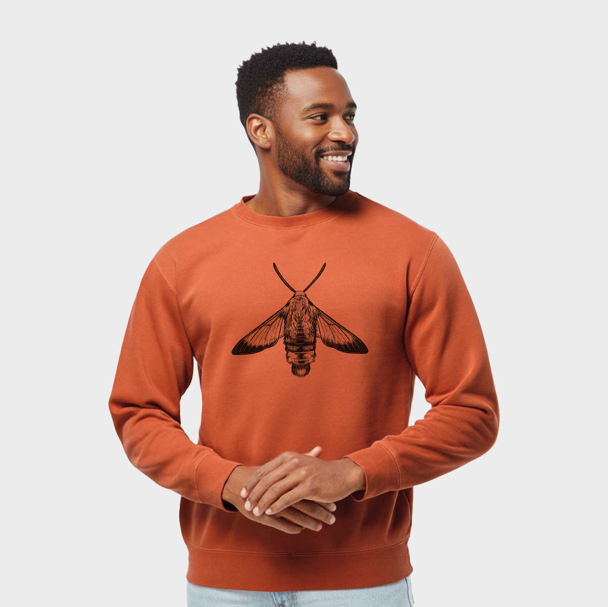 Model wearing an amber orange pigment dyed crewneck sweatshirt printed with a detailed black ink illustration of a snowberry clearwing moth, seen from above.