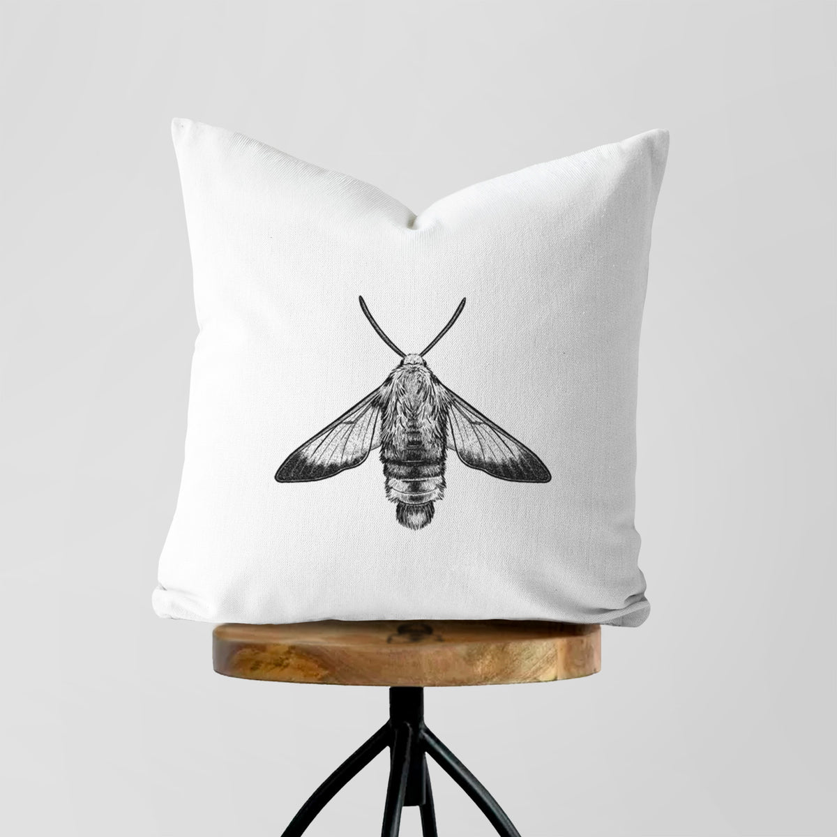 White pillow cover printed with a detailed black ink illustration of a snowberry clearwing moth, seen from above, on a wooden stool.