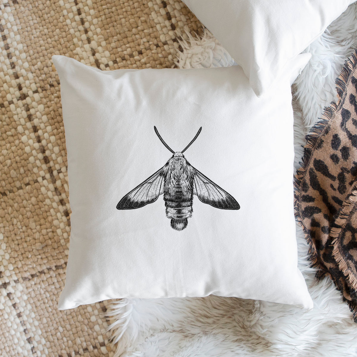 White pillow cover printed with a detailed black ink illustration of a snowberry clearwing moth, seen from above, on a woven chair.