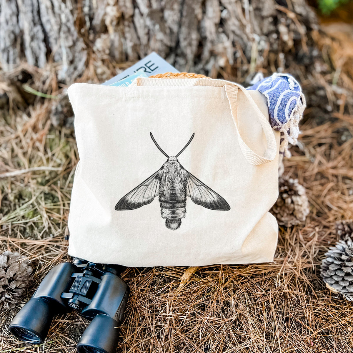 Canvas tote bag printed with a detailed black ink illustration of a snowberry clearwing moth, seen from above, resting on dry grass beside a pair of binoculars.