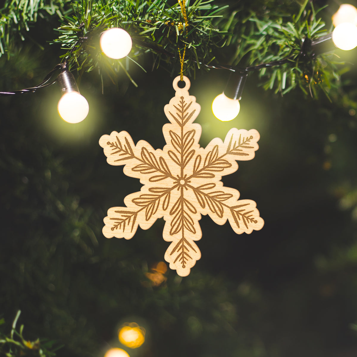 Wooden ornament featuring an engraved snowflake design with evergreen branches on each point, on a knit blanket with pine cones.