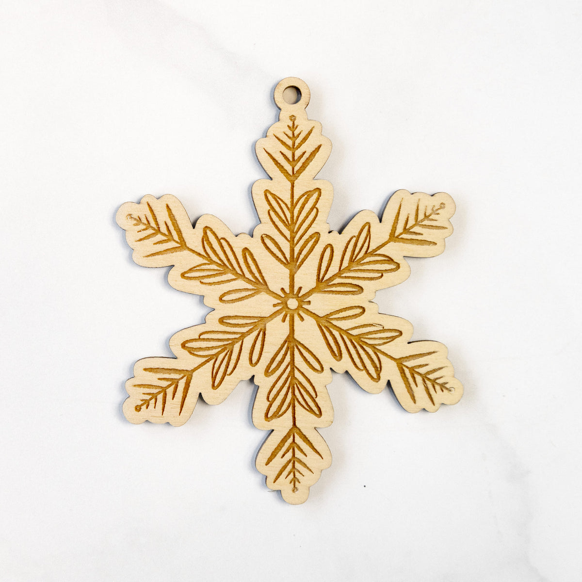 Wooden ornament featuring an engraved snowflake design with evergreen branches on each point, on a marble surface.