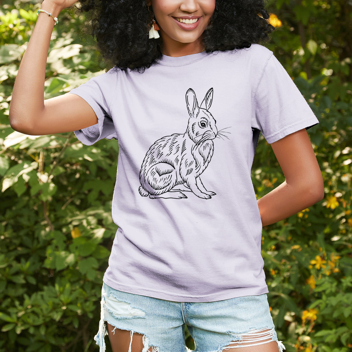 Woman wearing a Comfort Colors t-shirt printed with a detailed black ink illustration of a snowshoe hare sitting upright, outdoors.
