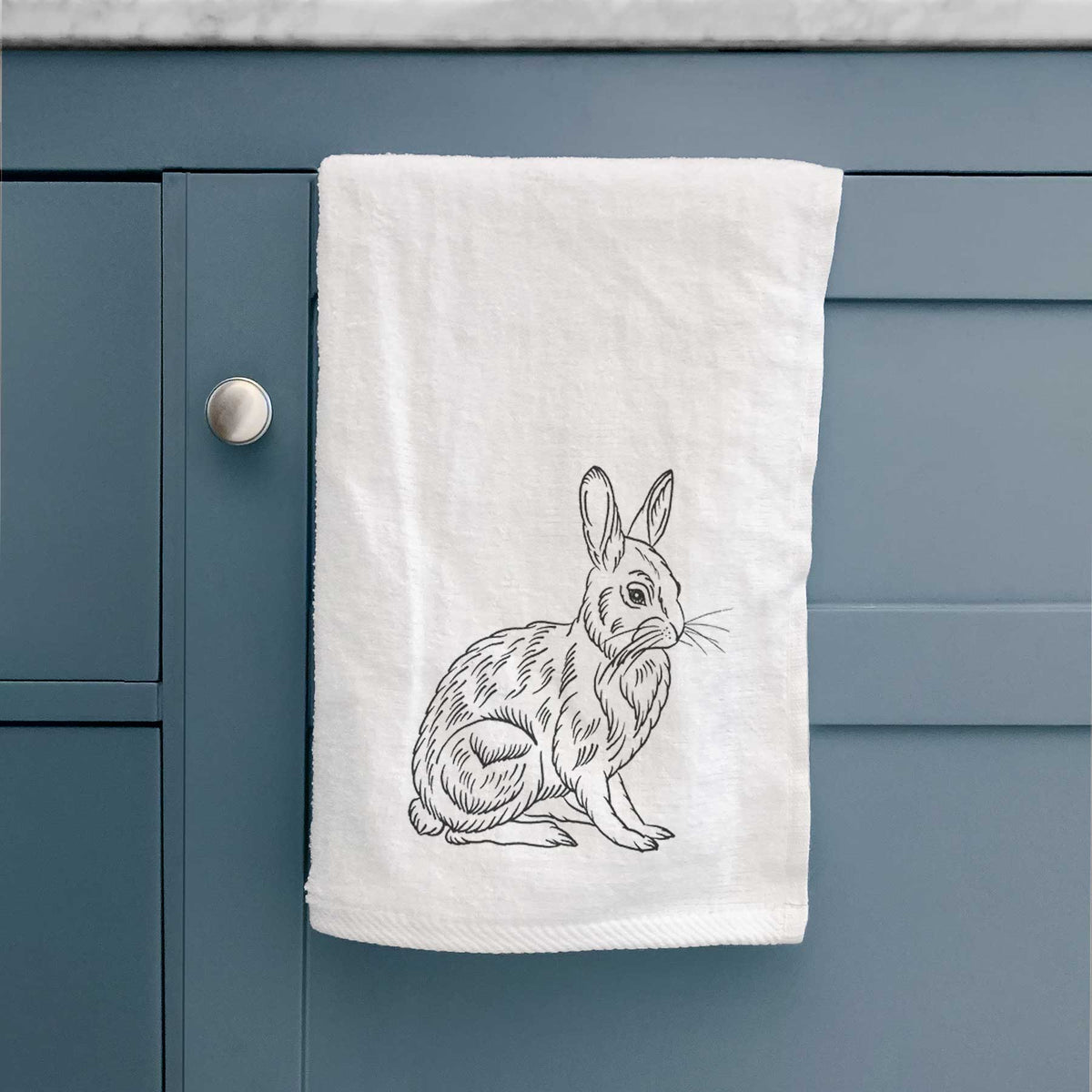 White hand towel printed with a detailed black ink illustration of a snowshoe hare sitting upright, hanging on a cabinet door.