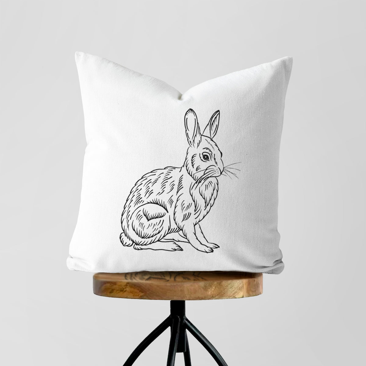 White pillow cover printed with a detailed black ink illustration of a snowshoe hare sitting upright, on a wooden stool.