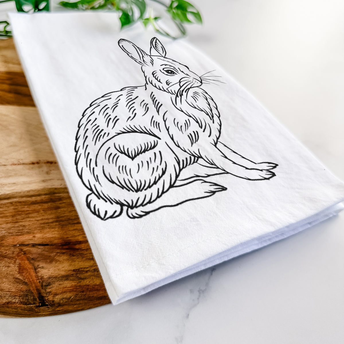 Close-up of a folded white tea towel on a wooden board, showing a detailed black ink illustration of a snowshoe hare sitting upright.