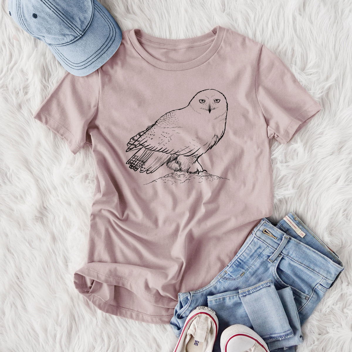 Women's relaxed fit cotton t-shirt printed with a detailed black ink illustration of a snowy owl perched on a snowy mound, styled flat on a white faux fur rug with jeans.