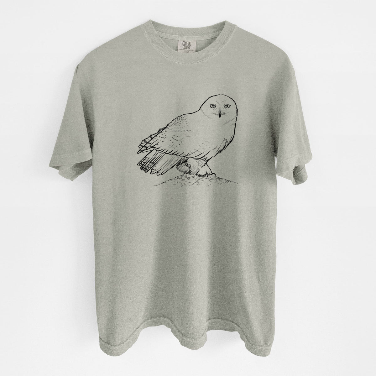 Sandstone beige Comfort Colors t-shirt laid flat, printed with a detailed black ink illustration of a snowy owl perched on a snowy mound.
