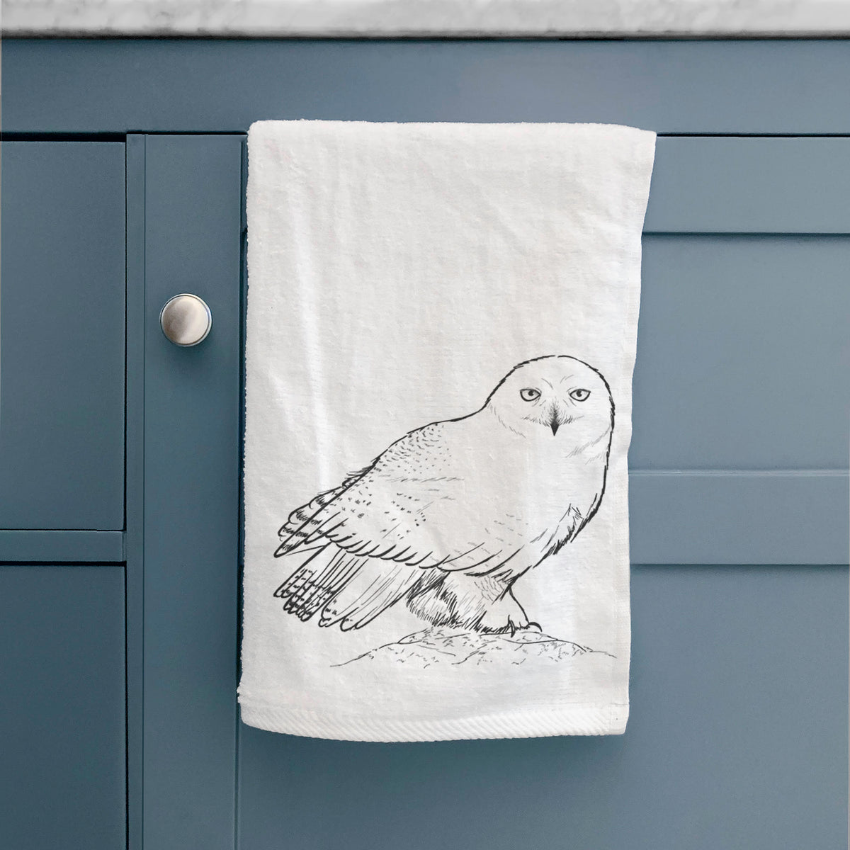 White hand towel printed with a detailed black ink illustration of a snowy owl perched on a snowy mound, hanging on a cabinet door.