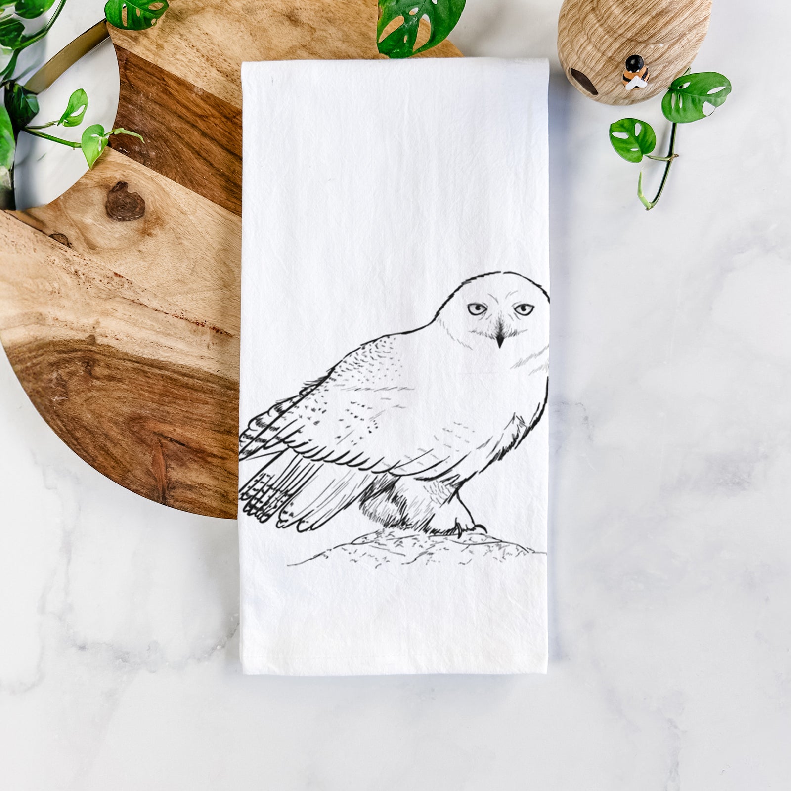 White cotton tea towel printed with a detailed black ink illustration of a snowy owl perched on a snowy mound, on a marble counter beside a round wooden cutting board and greenery.