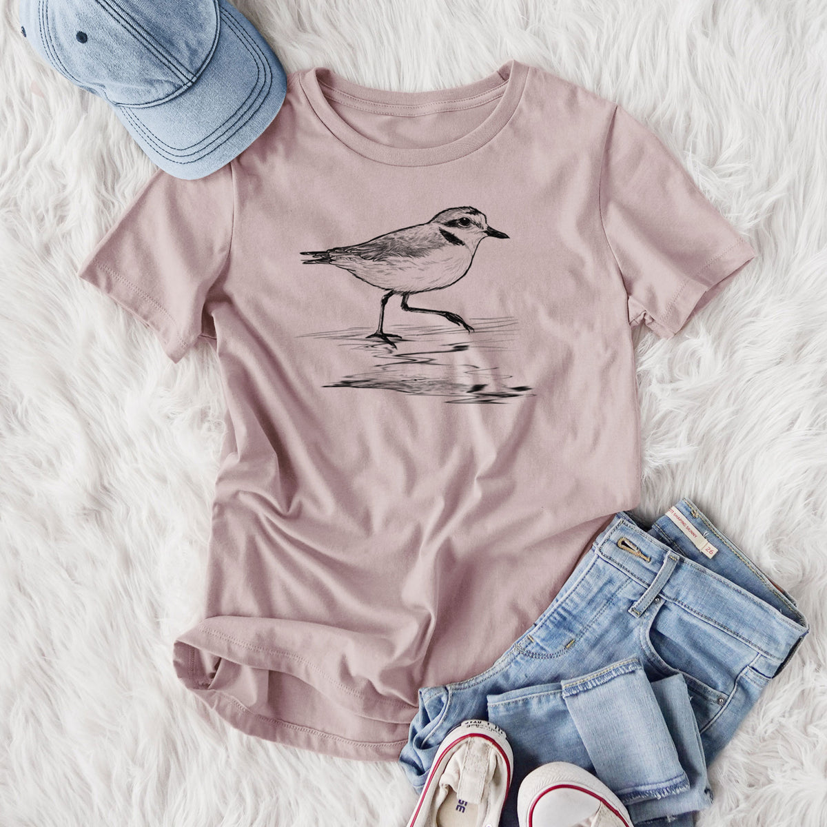 Women's relaxed fit cotton t-shirt printed with a black ink sketch of a snowy plover running along the wet shoreline, styled flat on a white faux fur rug with jeans.
