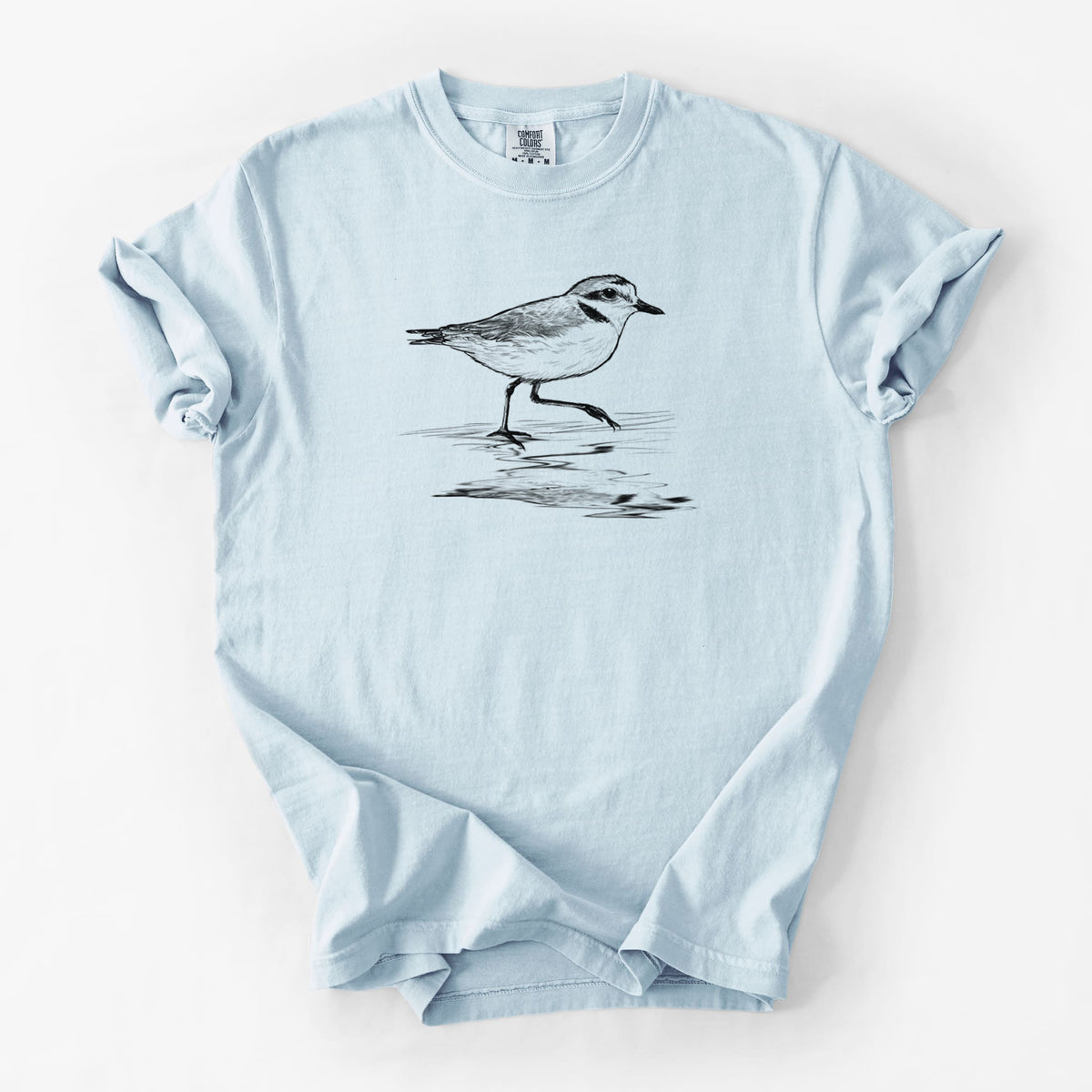 Chambray blue Comfort Colors t-shirt laid flat, printed with a black ink sketch of a snowy plover running along the wet shoreline.