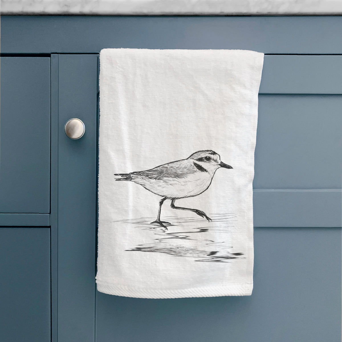 White hand towel printed with a black ink sketch of a snowy plover running along the wet shoreline, hanging on a cabinet door.