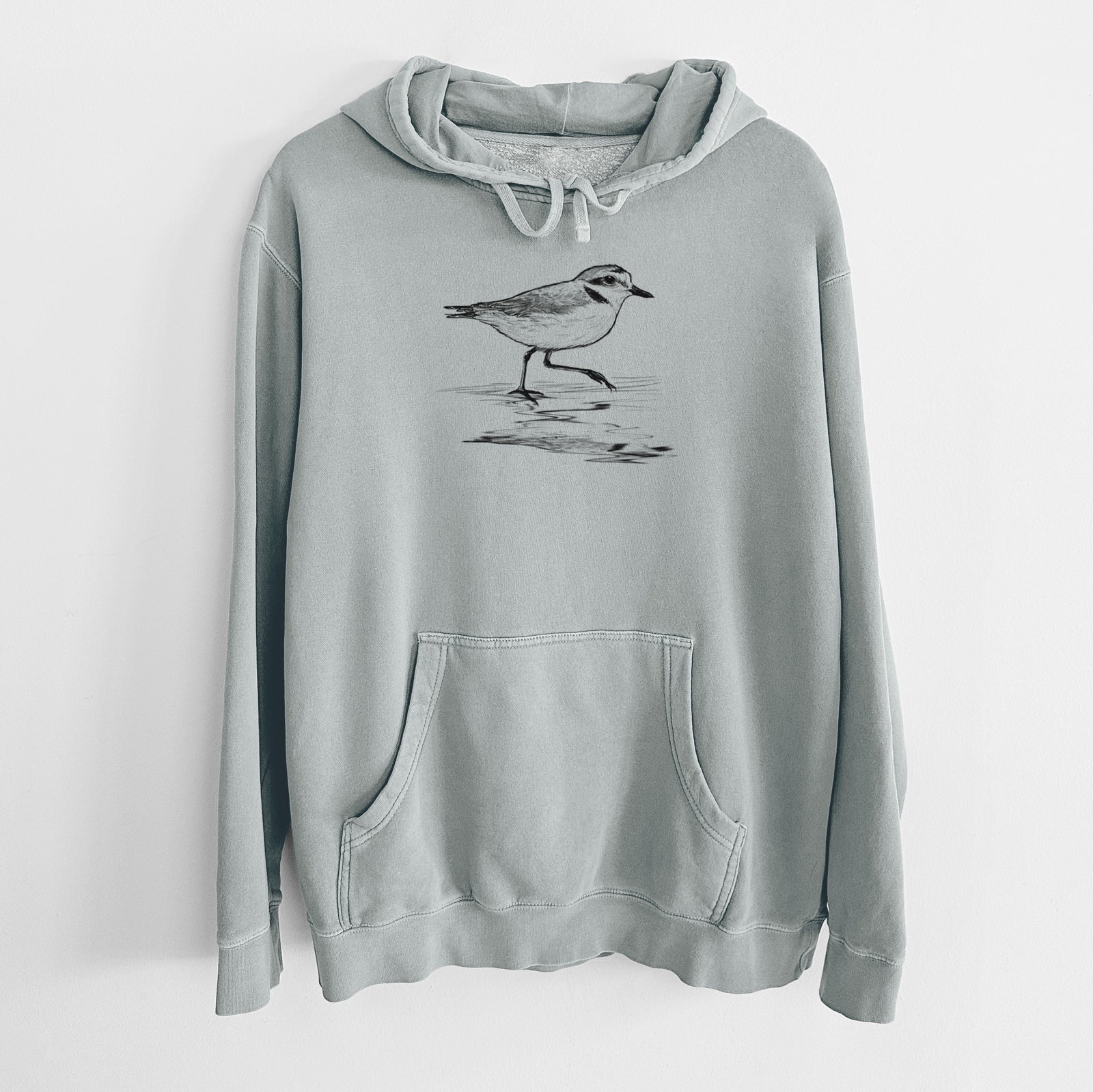 Sage green pigment dyed hoodie laid flat, printed with a black ink sketch of a snowy plover running along the wet shoreline.