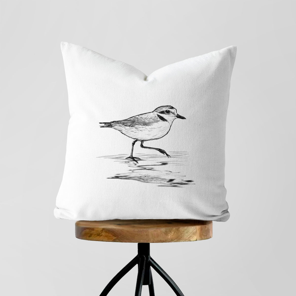 White pillow cover printed with a black ink sketch of a snowy plover running along the wet shoreline, on a wooden stool.