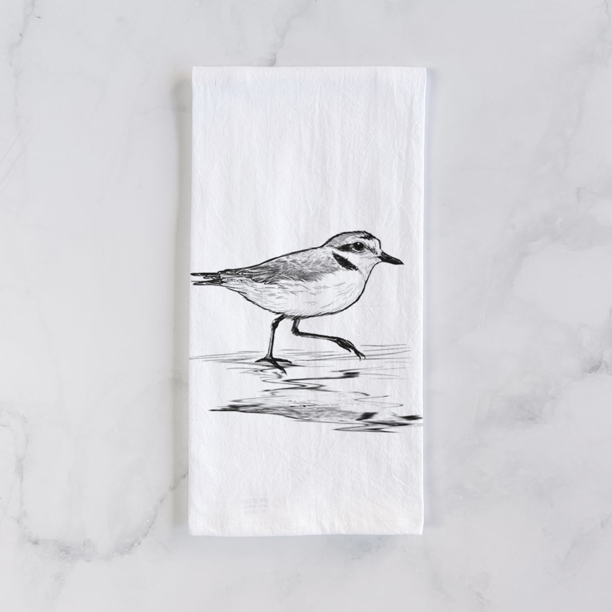 White cotton tea towel laid flat on marble, printed with a black ink sketch of a snowy plover running along the wet shoreline.