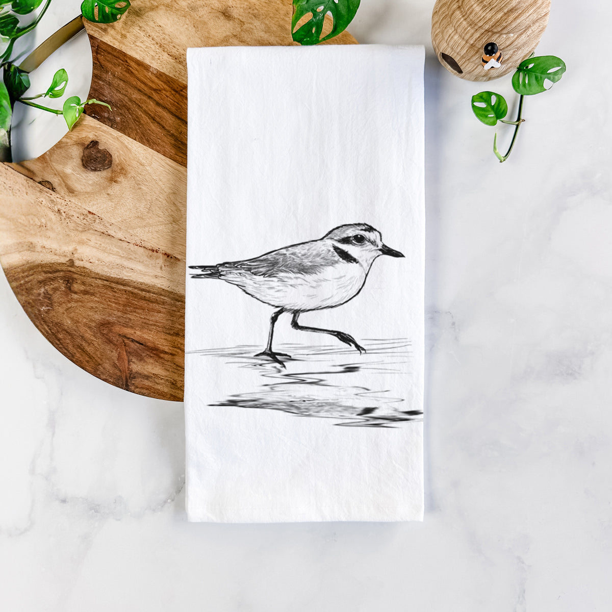 White cotton tea towel printed with a black ink sketch of a snowy plover running along the wet shoreline, on a marble counter beside a round wooden cutting board and greenery.