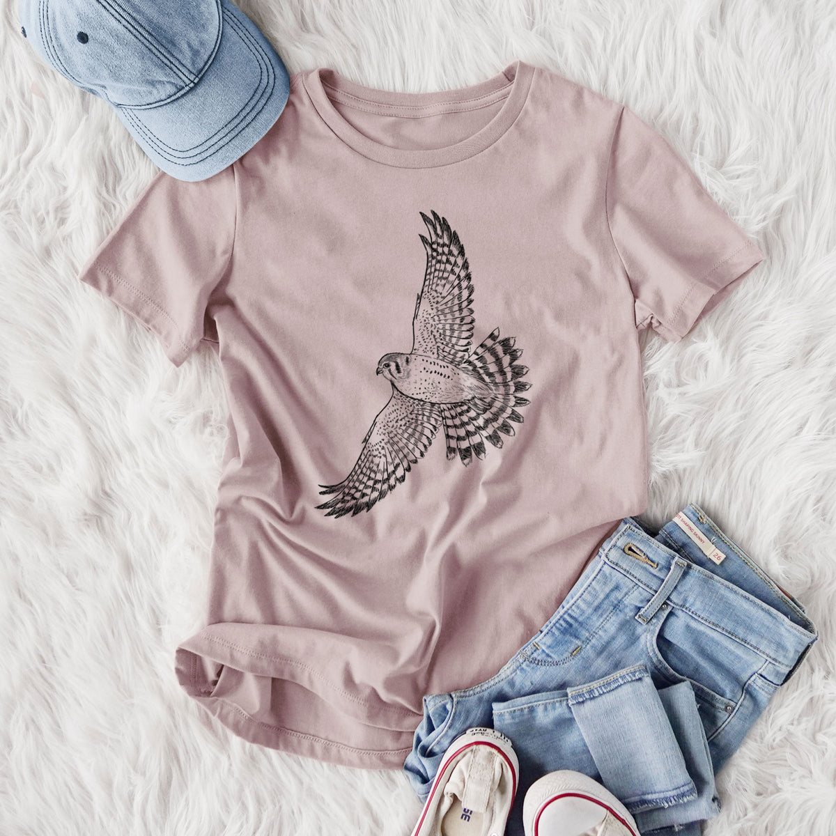 Women's relaxed fit cotton t-shirt printed with a detailed black ink illustration of an American kestrel in flight with its wings raised, styled flat on a white faux fur rug with jeans.