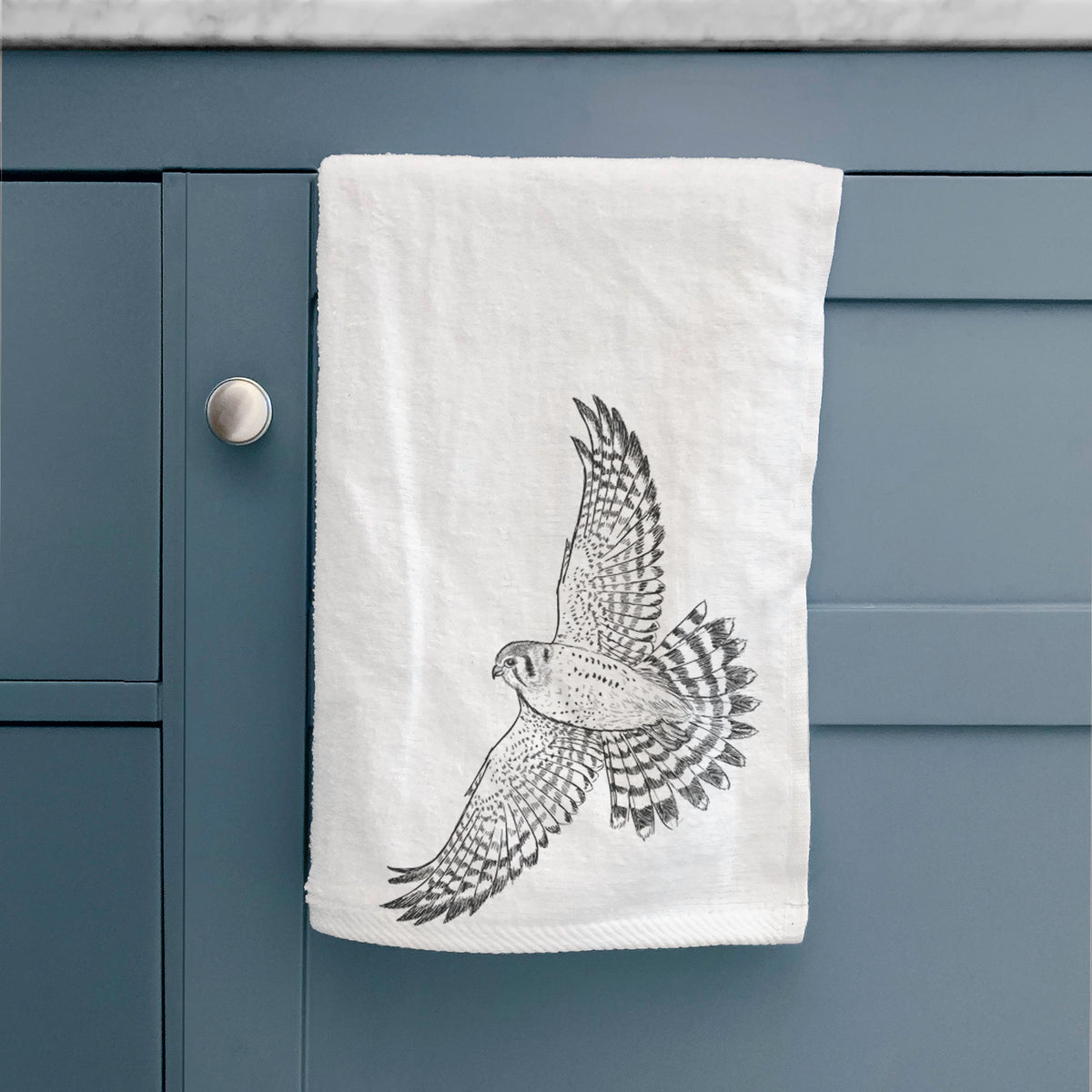 White hand towel printed with a detailed black ink illustration of an American kestrel in flight with its wings raised, hanging on a cabinet door.