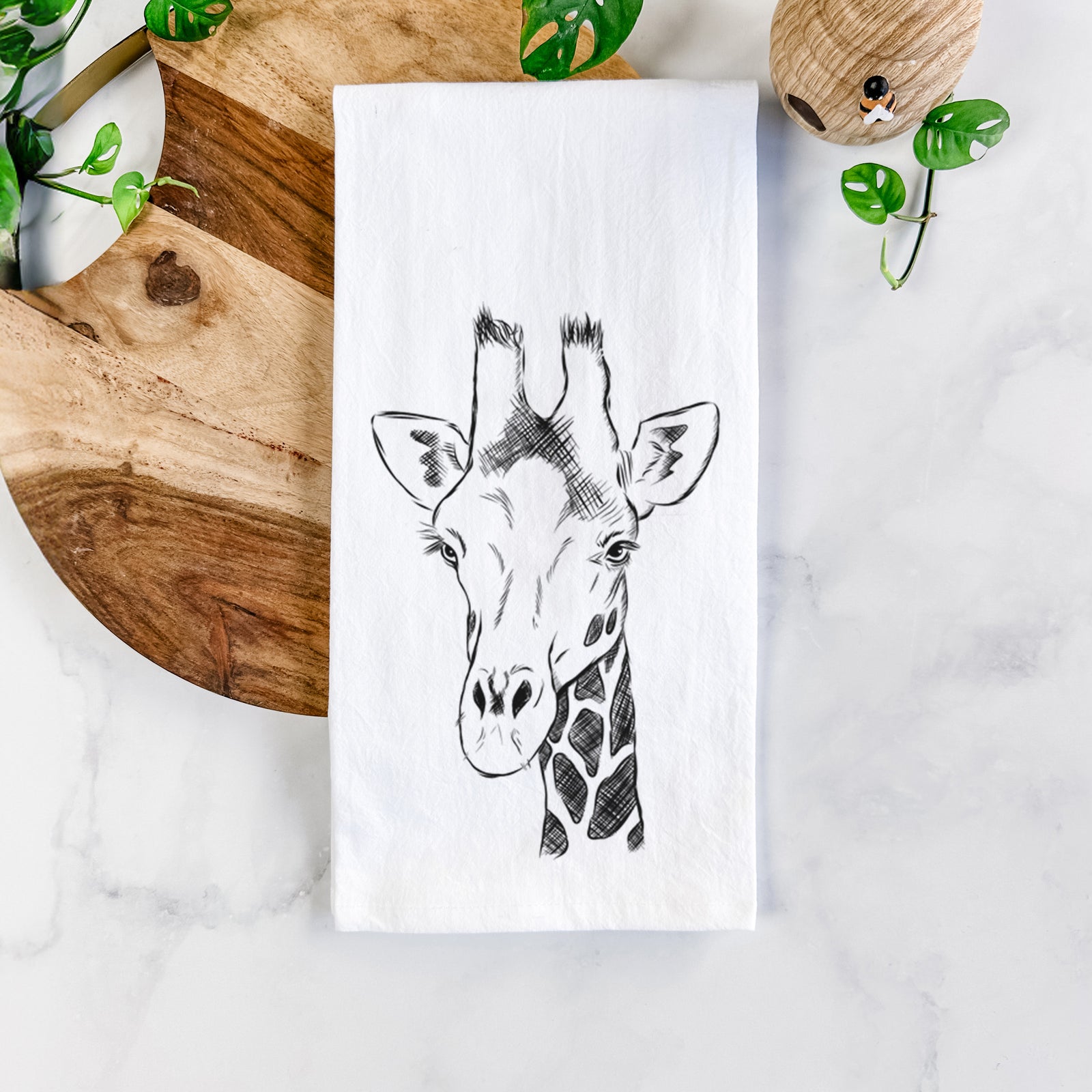White cotton tea towel printed with a black ink portrait of a southern giraffe's head and neck, on a marble counter beside a round wooden cutting board and greenery.