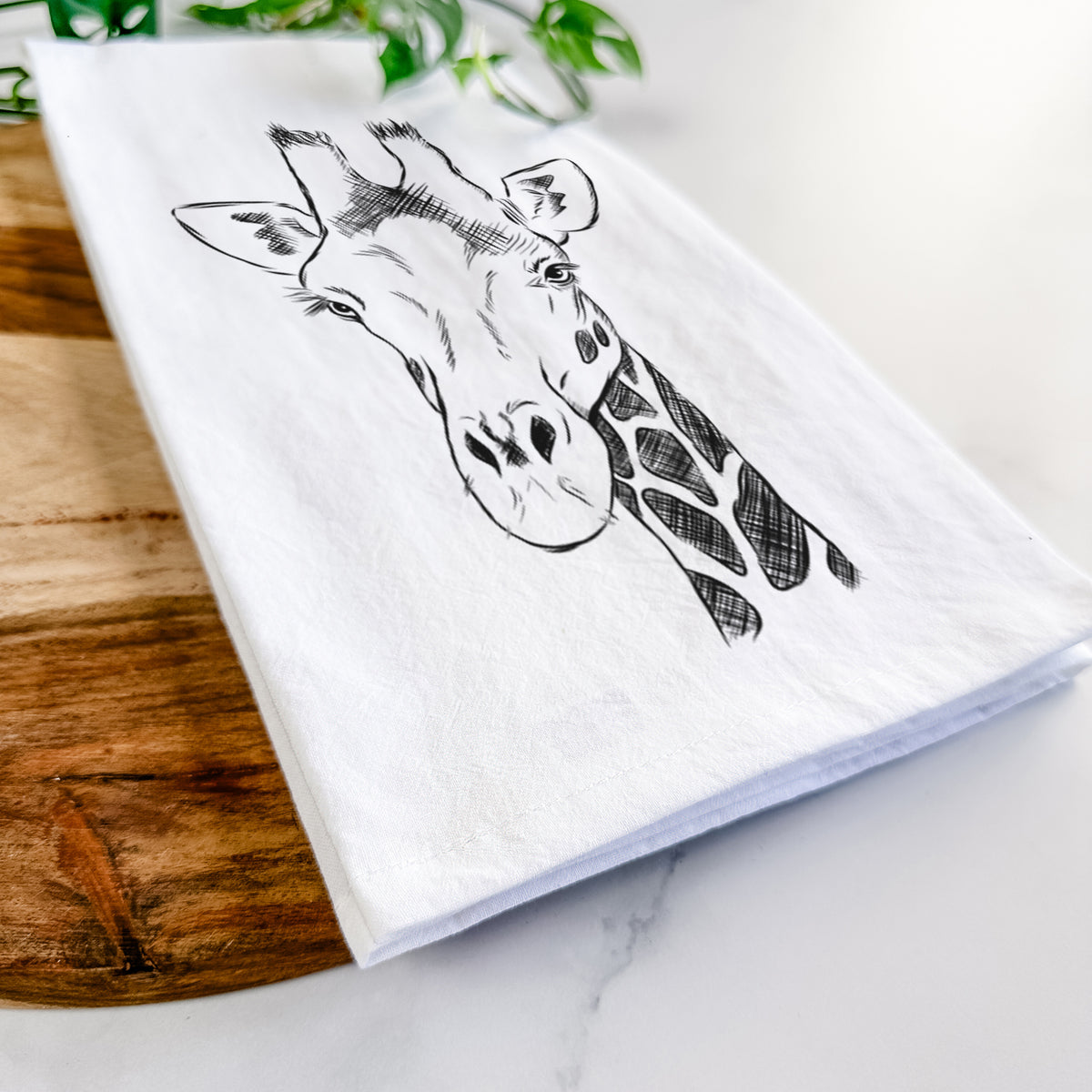 Close-up of a folded white tea towel on a wooden board, showing a black ink portrait of a southern giraffe's head and neck.