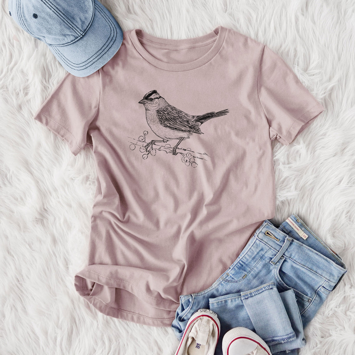 Women's relaxed fit cotton t-shirt printed with a detailed black ink sketch of a white-crowned sparrow perched on a branch, styled flat on a white faux fur rug with jeans.