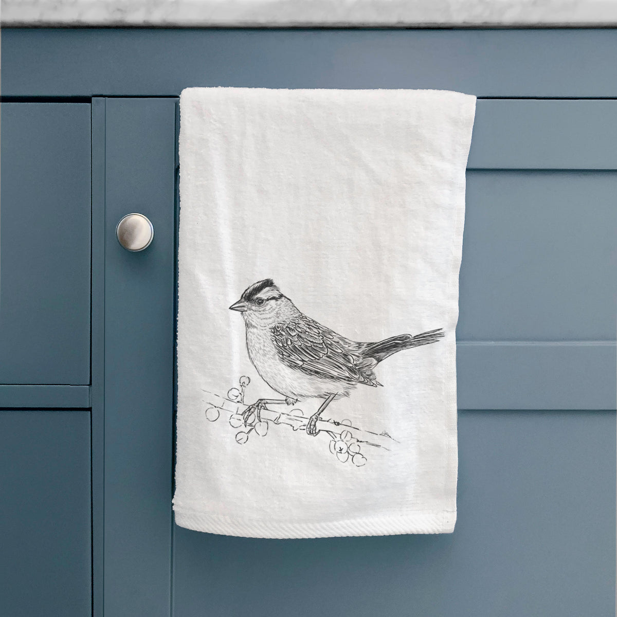 White hand towel printed with a detailed black ink sketch of a white-crowned sparrow perched on a branch, hanging on a cabinet door.